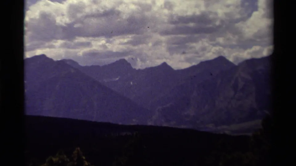 Zoomed 1979 8mm view of rugged Canadian mountain ridges, mottled clouds, and a dark forested foreground band.