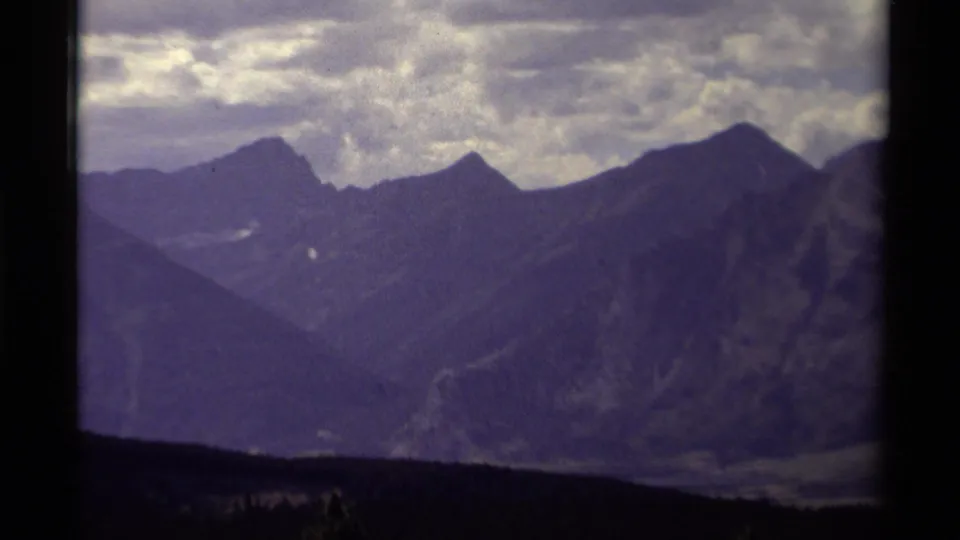 Tight 8mm home-movie view of Canadian mountain peaks and hazy ridges beneath a textured cloud layer, with visible purple color shift.