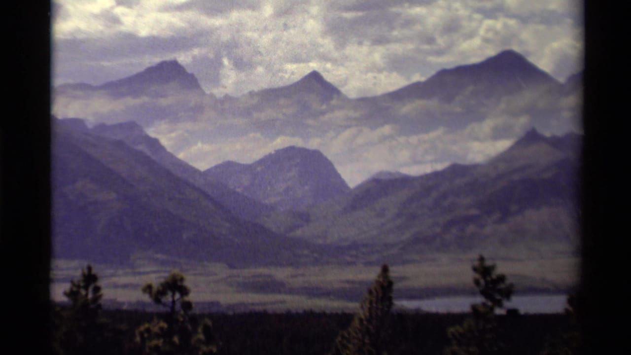 1979 Canadian 8mm frame showing a mountain valley, distant peaks, foreground trees, and a pale lake or river.