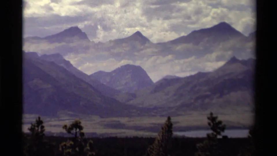 1979 Canadian 8mm frame showing a mountain valley, distant peaks, foreground trees, and a pale lake or river.