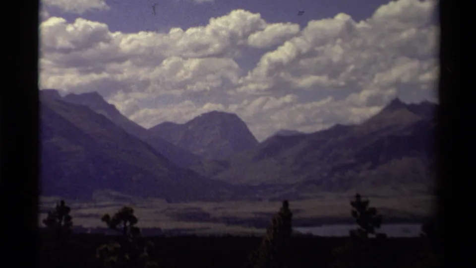 1979 Canadian 8mm landscape frame with a shadowed valley, layered mountains, large clouds, and dark foreground trees.