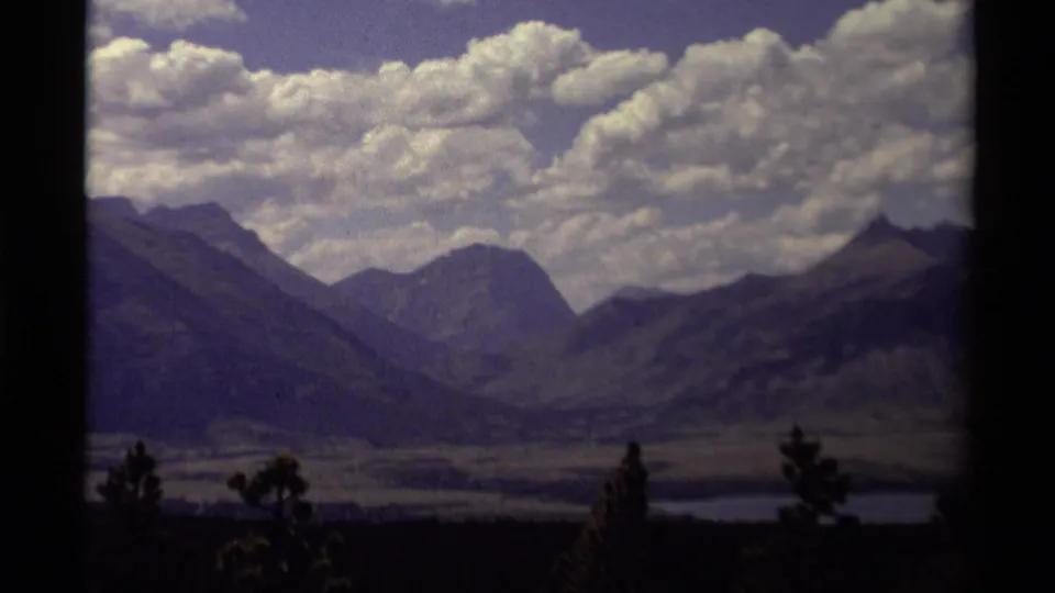 1979 8mm Canadian mountain valley with layered peaks, cloud-filled sky, and a dark foreground frame suggesting a vehicle or window view.