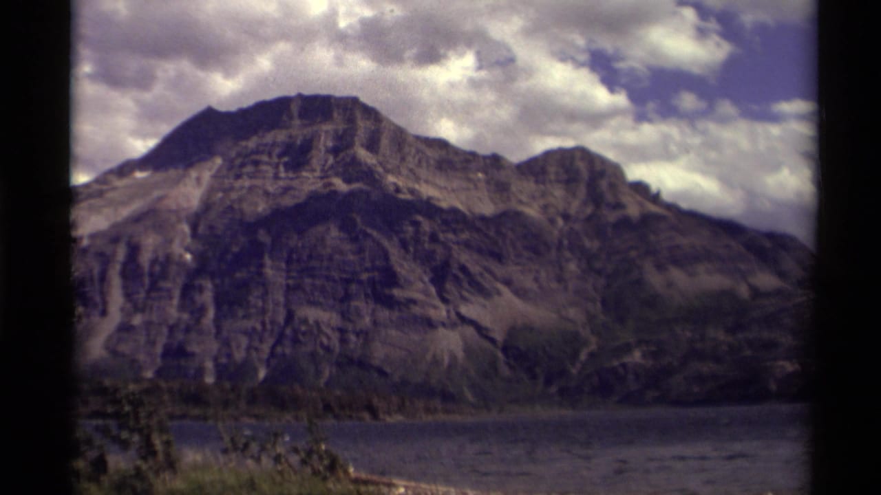 Rocky Canadian mountain rising above a calm lake under broken white clouds, recorded with soft 8mm color and dark edge framing.