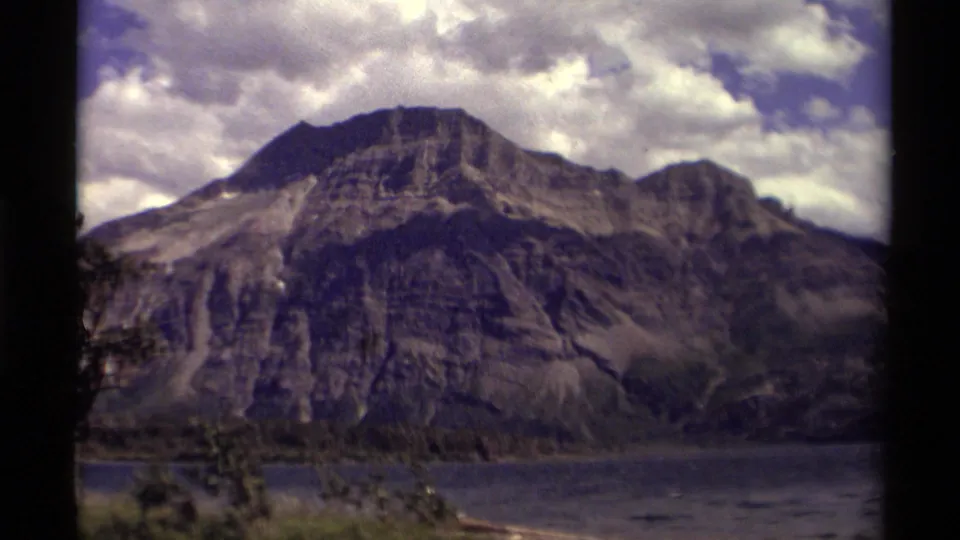 1979 Canadian mountain landscape with a rocky peak, lake foreground, sparse trees, and saturated 8mm color under cloudy sky.