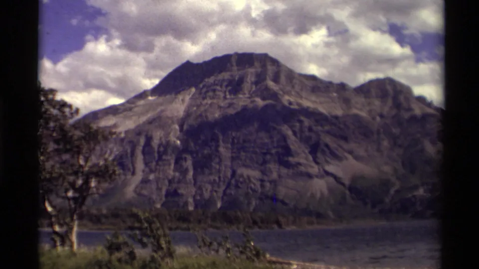 Canadian mountain and lake scene from 1979 home-movie footage, with foreground tree branches, cloudy sky, and soft 8mm texture.