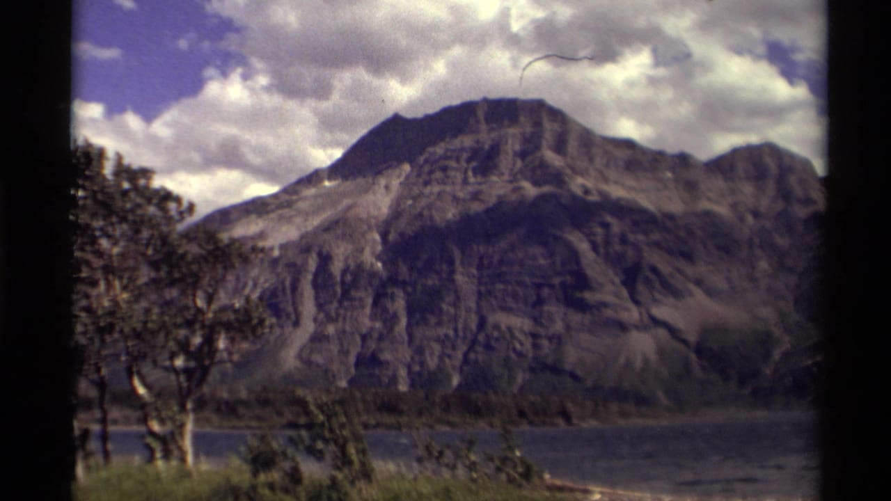 Rocky mountain above a lake, with trees at left and clouds overhead.