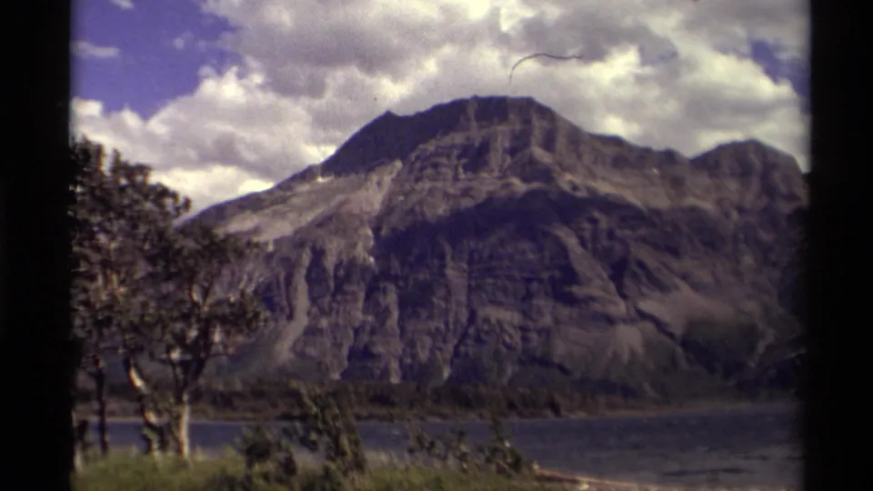 1979 Canadian 8mm frame showing a rocky mountain rising beyond a lake, with shoreline trees and a cloudy sky.