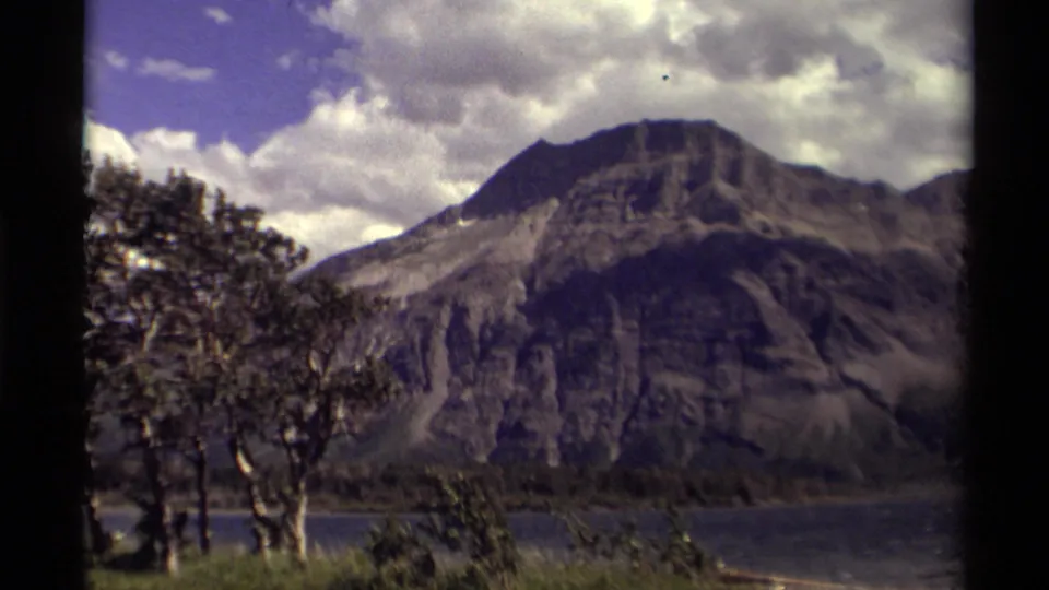 1979 Canadian 8mm frame of a rugged mountain across a lake, partly framed by foreground trees and bright clouds.