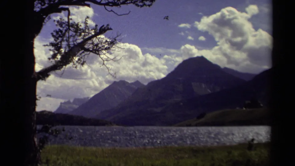 1979 Canadian 8mm frame showing darker mountain peaks beyond a lake, with overhanging branches and sunlit clouds.