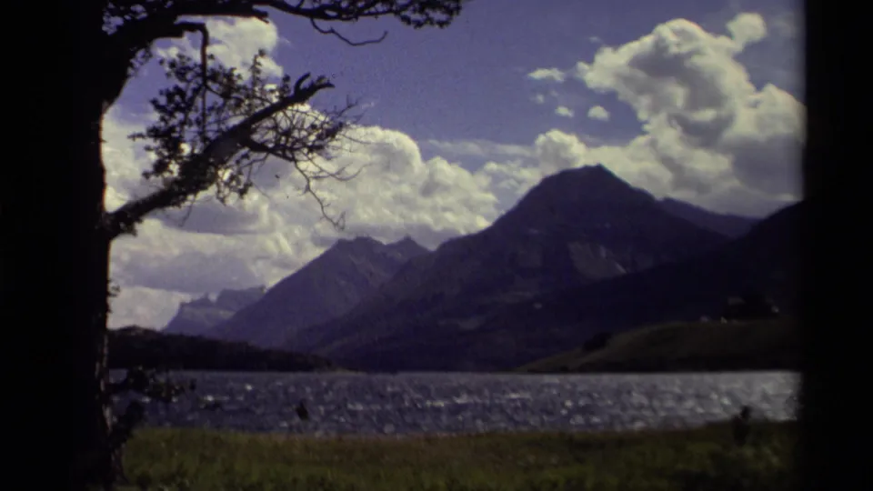 1979 Canadian 8mm frame showing a mountain beyond a lake, with dark foreground trees and bright clouds overhead.