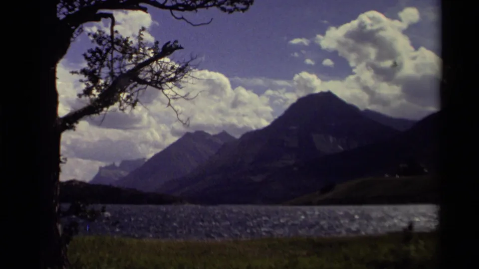 1979 Canadian 8mm frame of a calm lake and mountain ridge, partly framed by a dark tree trunk and branches.