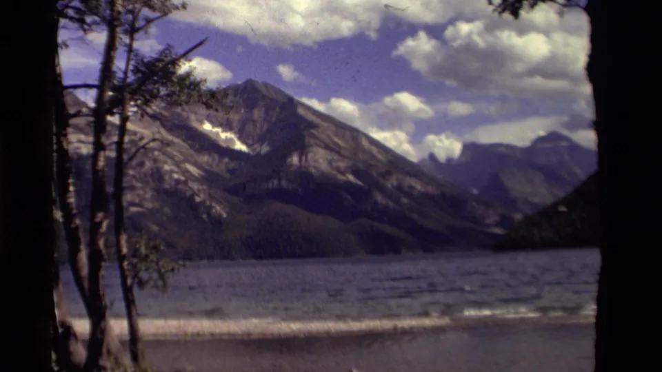 1979 Canadian 8mm frame showing a broad lake before rugged mountains, with slender trees along the left edge.