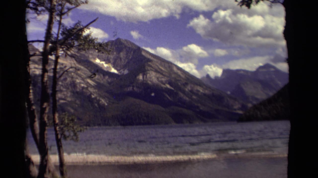 Lake and distant mountains framed by dark trees under a partly cloudy sky.