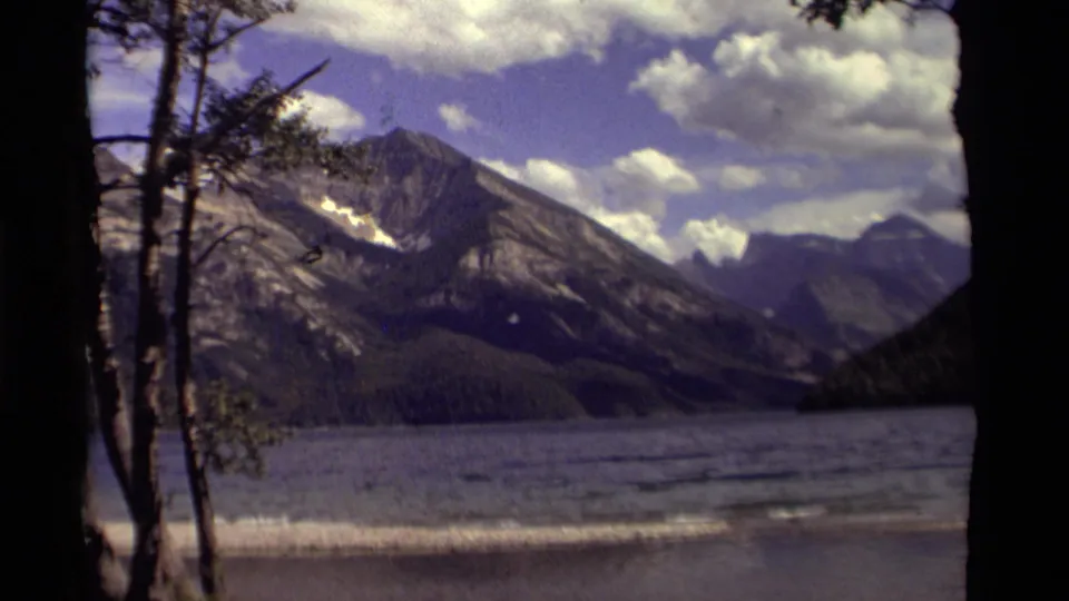 1979 Canadian 8mm view of a lake, forested slopes, and distant mountains framed by dark tree silhouettes.