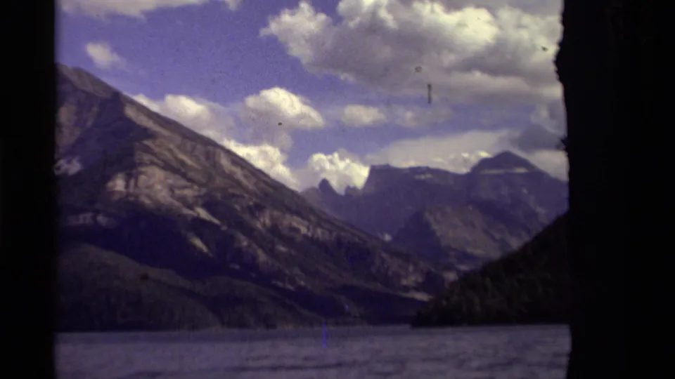 A closer 8mm frame shows rugged Canadian mountains rising beyond the lake, with forested lower slopes and cloud cover above.