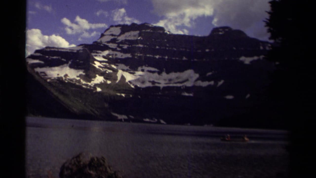 Small boat on a mountain lake beneath snow-dappled peaks and cloudy sky.