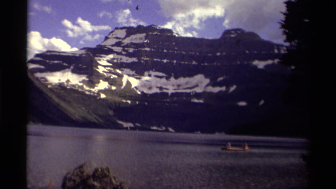 Small boat moving across calm lake water with snowy mountain peaks behind it.