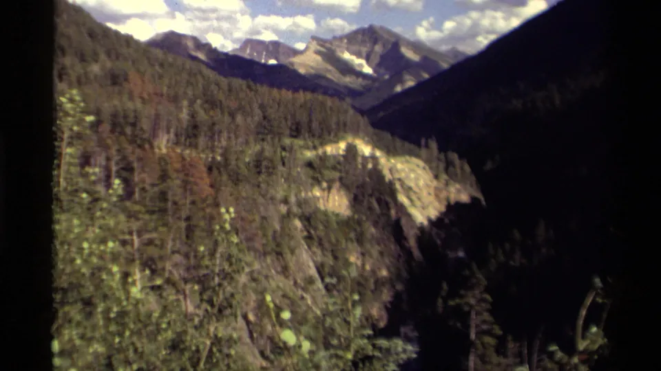 1979 Canadian 8mm view of a forested valley leading toward rugged mountain peaks under scattered clouds.
