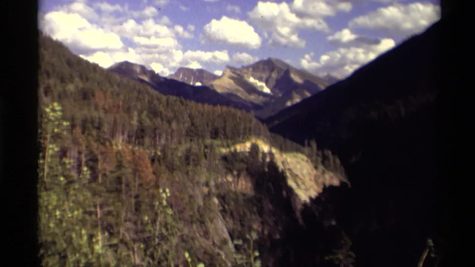 Canadian 8mm travel frame with evergreen-covered slopes, a mountain valley, and pale clouds over distant peaks.