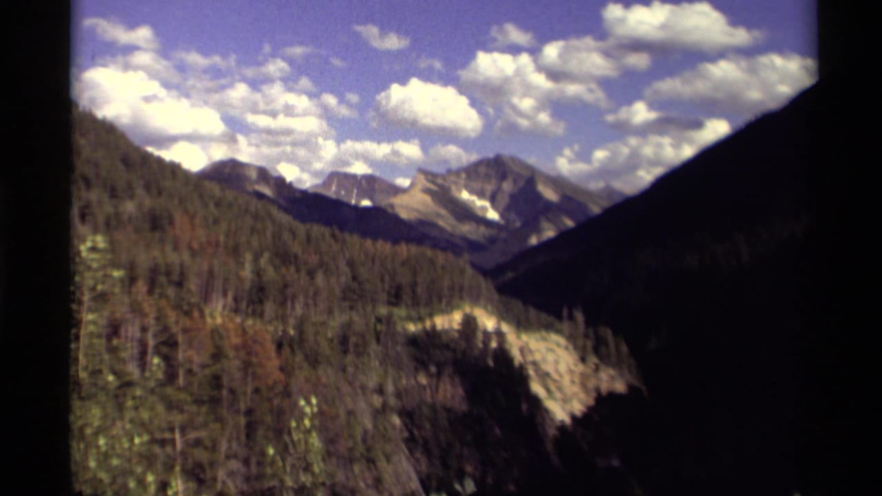 Forested mountain valley with distant peaks under scattered white clouds.