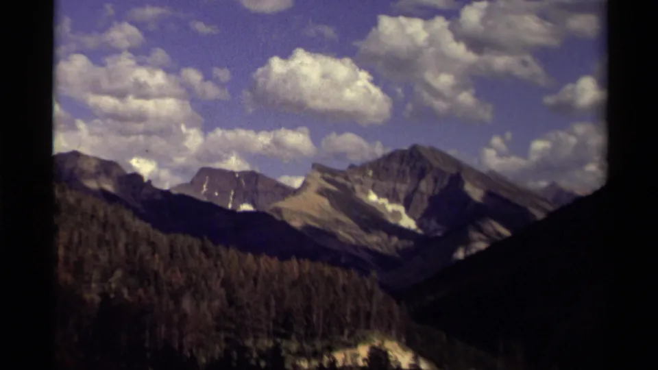 1979 Canada 8mm frame of a distant mountain range, evergreen foreground, and scattered clouds, with dark film borders visible.