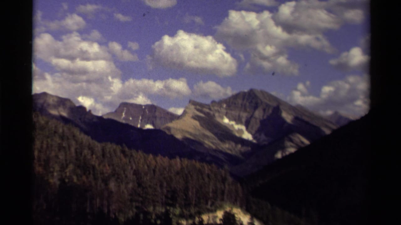 Distant rocky mountains above evergreen forest under a cloud-filled blue sky.