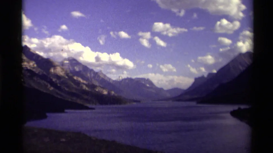 1979 Canada 8mm view of a dark alpine lake stretching between mountain slopes under scattered white clouds.