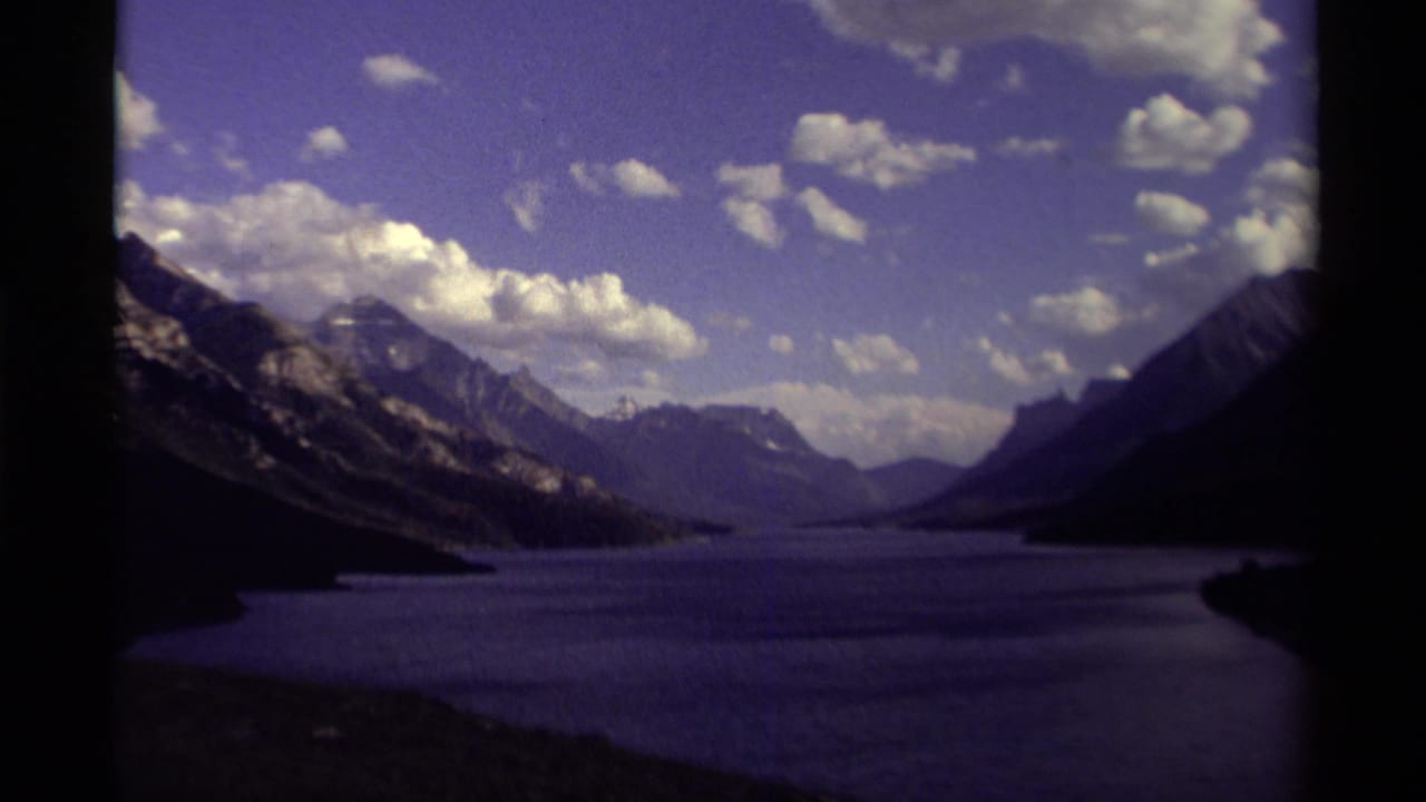 1979 8mm home-movie view of a Canadian mountain lake framed by rugged slopes, distant peaks, clouds, and dark film borders.