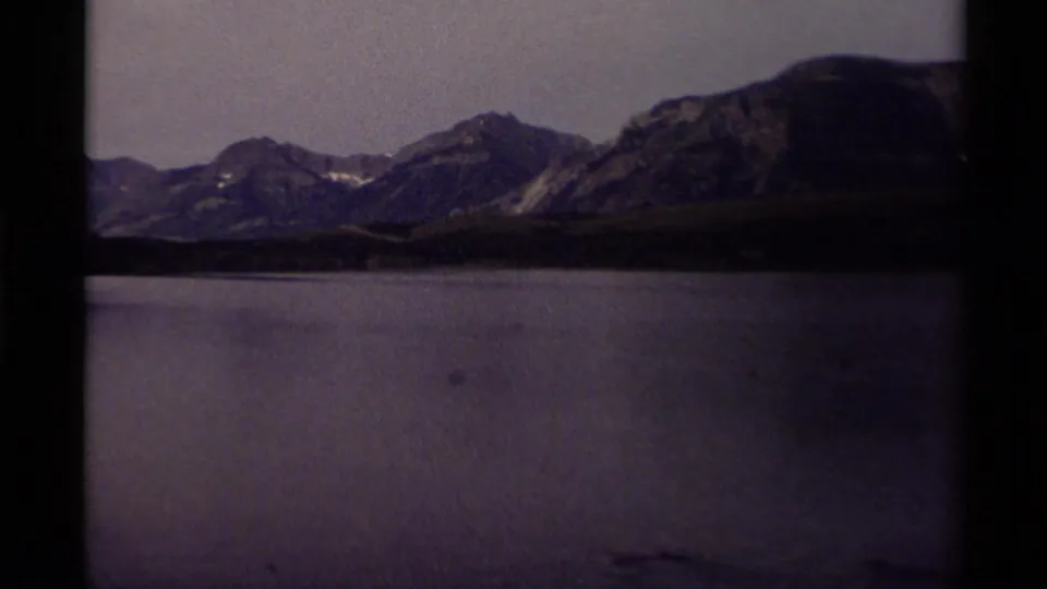 1979 Canadian lake and mountain scene with low forest along the shore, layered ridges, muted sky, and visible home-movie grain.