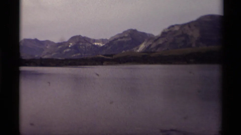 Canadian mountain lake view from 1979 8mm footage, showing calm water, rocky peaks, a forested bank, and analog color fading.