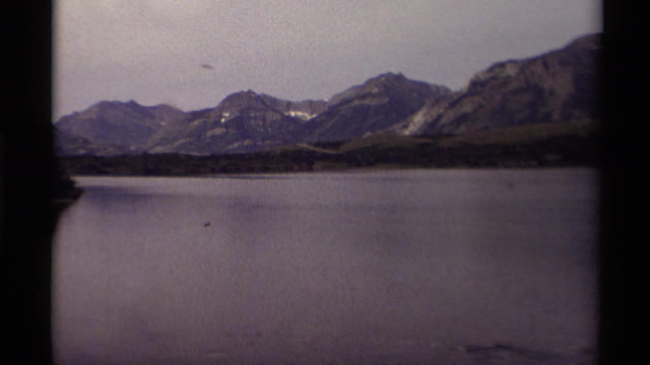 1979 8mm home-movie frame of a Canadian mountain lake with dark shore trees, distant ridges, overcast sky, and small birds visible on the water.