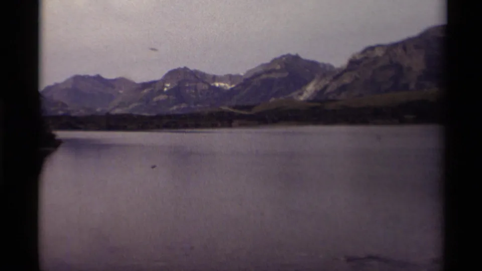 1979 8mm home-movie frame of a Canadian mountain lake with dark shore trees, distant ridges, overcast sky, and small birds visible on the water.