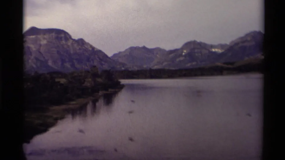 Late-1970s 8mm frame from Canada showing birds on a calm lake, a tree-lined point, layered mountains, and muted home-movie color.