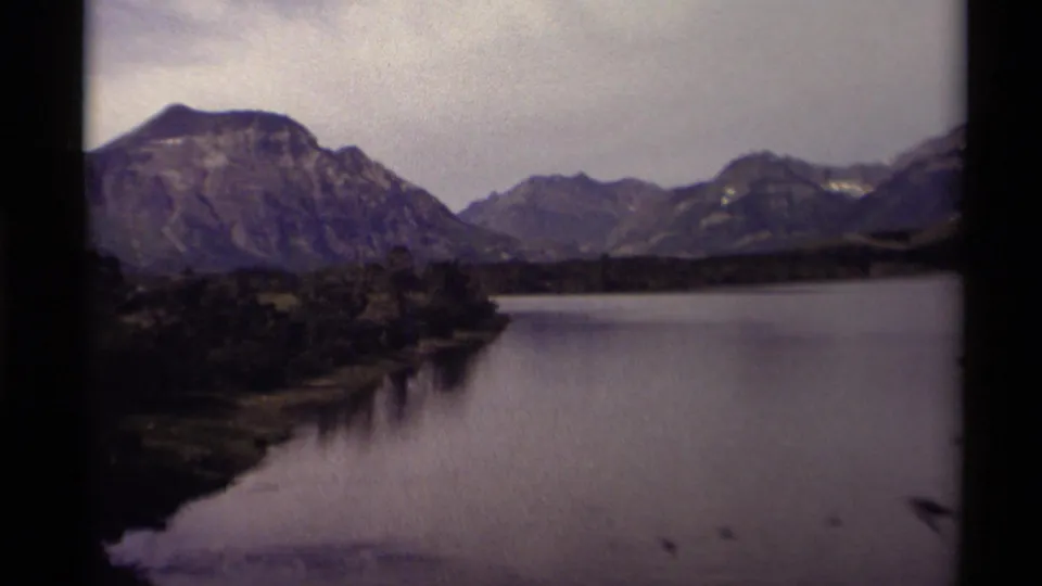 1979 8mm home-movie view of a calm Canadian lake beneath rugged mountains, with trees along the shore and dark film borders.