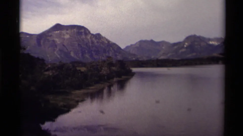 Canadian mountain lake scene on 8mm film, showing still water, forested shoreline, rugged peaks, and cloudy light.