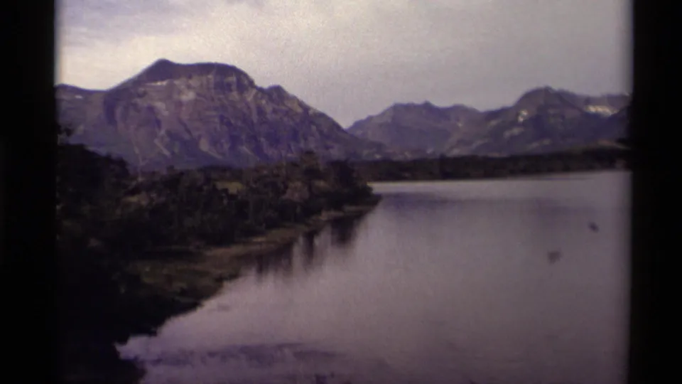 1979 Canadian 8mm landscape frame with a broad lake, dark wooded shoreline, distant mountains, and overcast sky.