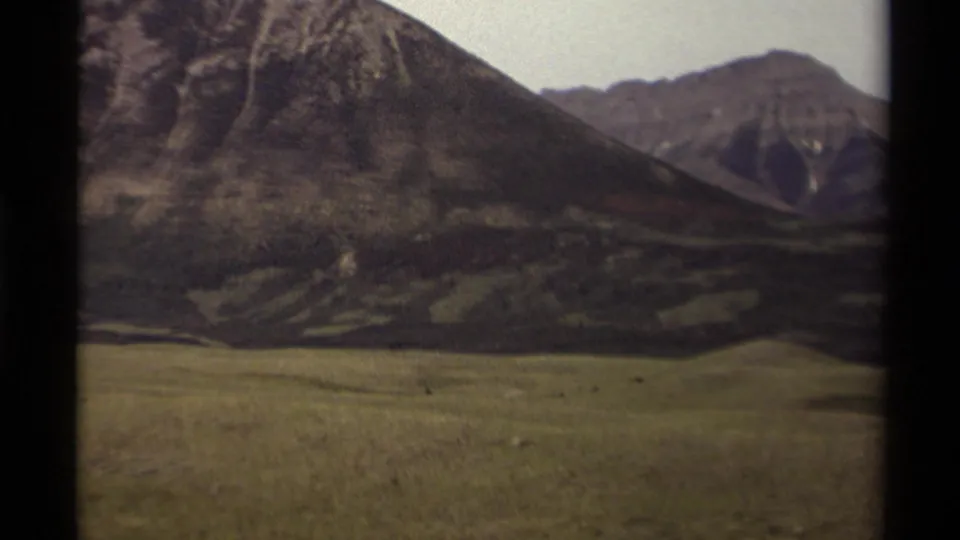 1979 Canadian home-movie view of rolling grassland below rugged mountain slopes and a clear, pale sky.