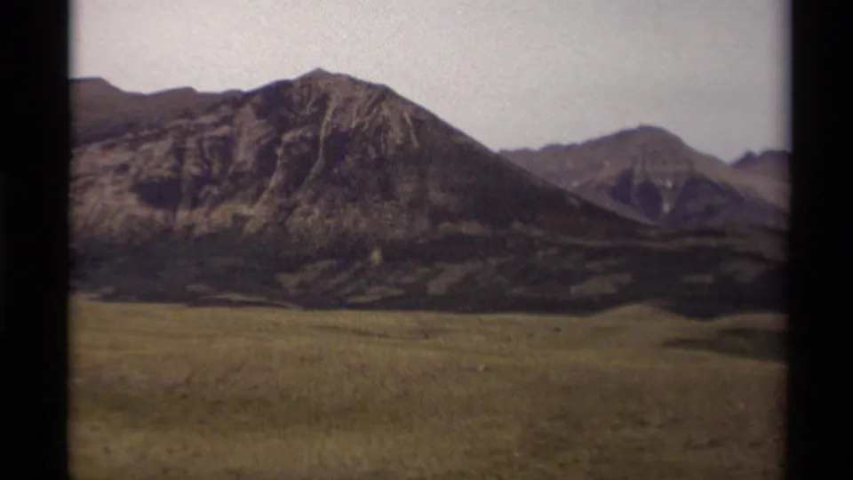 1979 Canadian 8mm frame centered on rugged mountain terrain rising behind a broad green field.