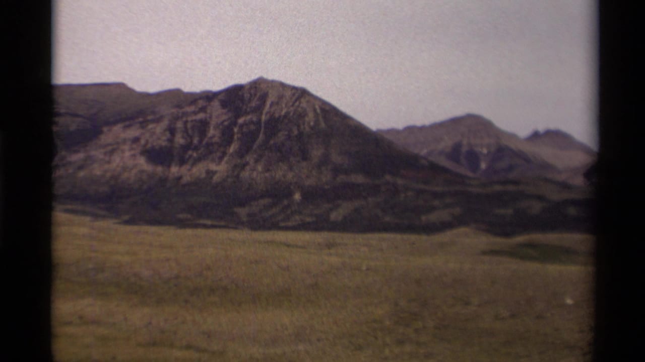 1979 Canadian home-movie frame showing rocky mountains and open grassland under a washed pale sky, bordered by dark film edges.