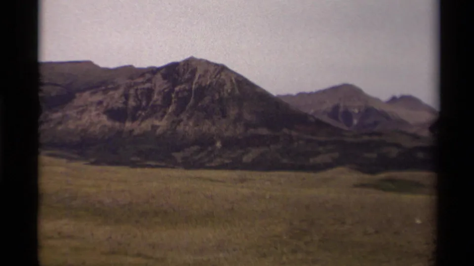 1979 Canadian home-movie frame showing rocky mountains and open grassland under a washed pale sky, bordered by dark film edges.