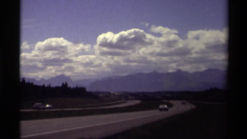 1979 Canadian Rockies highway view with small cars on a curving road, bright cloud banks, distant peaks, and dark foreground film exposure.