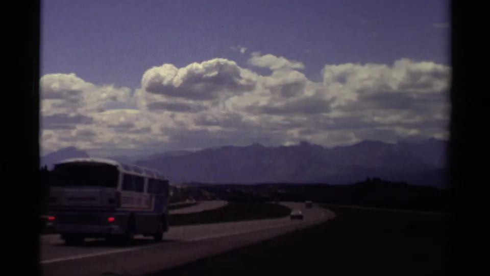 1979 Canadian Rockies road scene with a highway bus and cars moving along a curve under large clouds and distant mountain silhouettes.