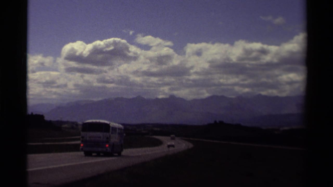 1979 Canadian 8mm road-trip frame showing a bus and cars on a curving highway, with foothills, distant mountains, and large clouds.