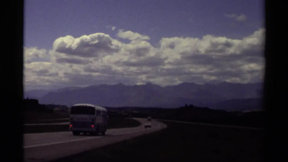 1979 Canadian 8mm road-trip frame showing a bus and cars on a curving highway, with foothills, distant mountains, and large clouds.