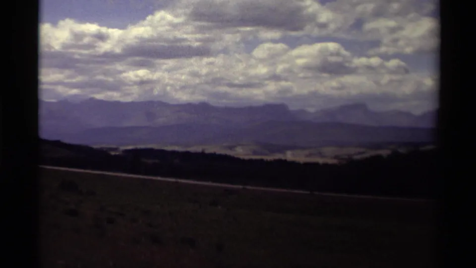 1979 Canadian 8mm road-trip frame emphasizing open foothills, a distant mountain range, broad clouds, and the dark edge of the film frame.