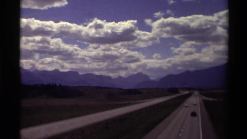 1979 Canadian 8mm frame of a multi-lane highway receding toward distant mountains under large clouds.