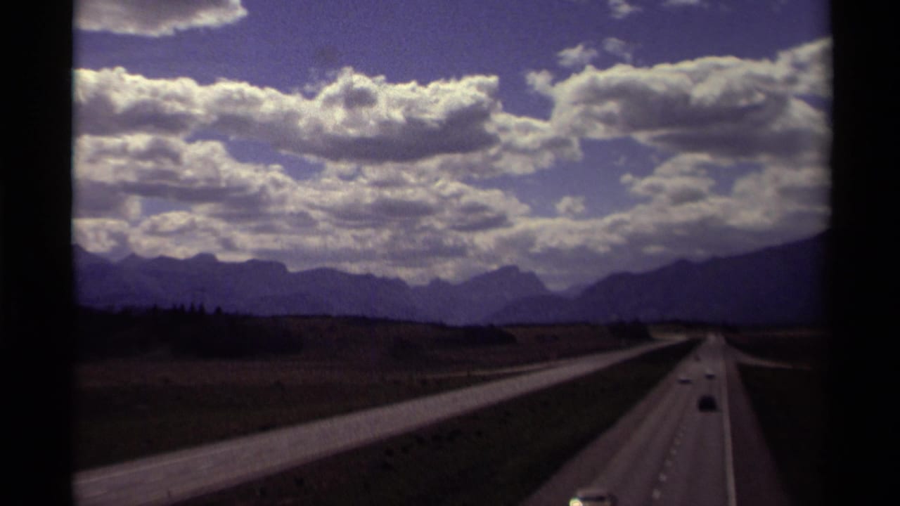 Cars travel along a divided highway toward distant mountains under a cloudy sky.