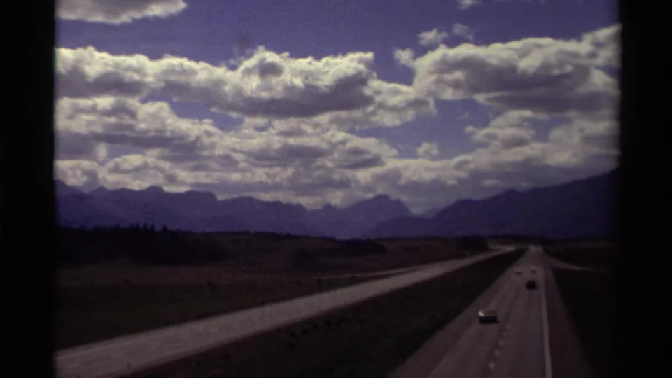 Canadian 8mm road view with cars on a multi-lane highway, open land, and mountains below a textured cloudy sky.