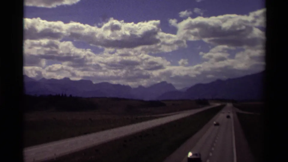 1979 Canadian home-movie frame showing cars on a divided highway with fields, trees, mountains, and heavy cloud cover.