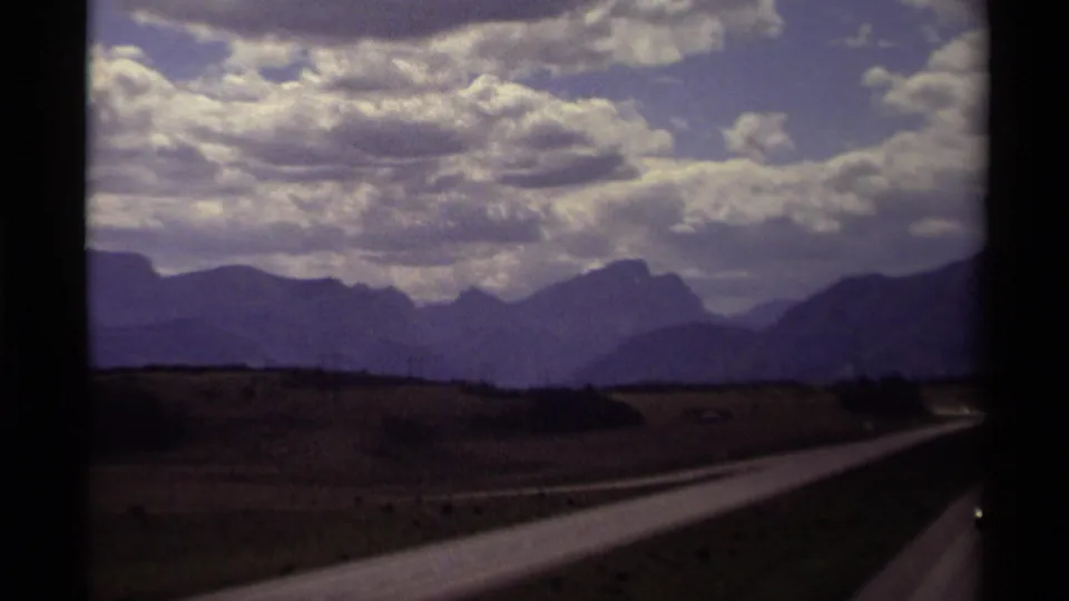 Canadian mountain landscape seen from a moving vehicle, with roadway, fields, and layered ridges under bright clouds.